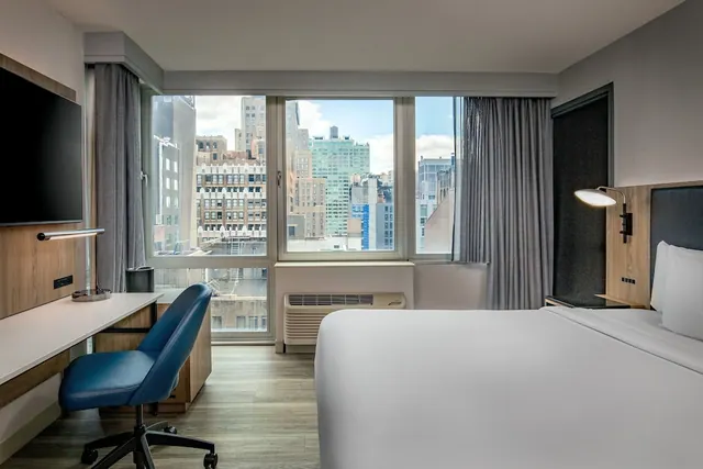 A bedroom with a king bed, a wall-mounted television, a desk with a blue chair, and large windows overlooking city buildings.