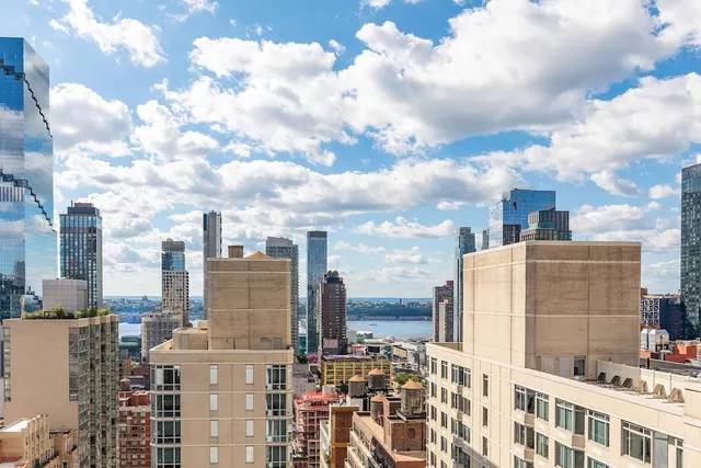 View from an upper floor across rooftops and high-rise towers toward a river under a partly cloudy blue sky.