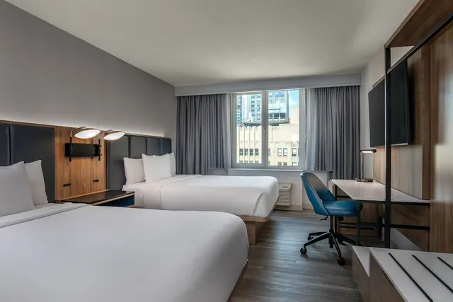 Two made beds face a desk with a blue chair and a wall-mounted TV, with a window showing city buildings outside.