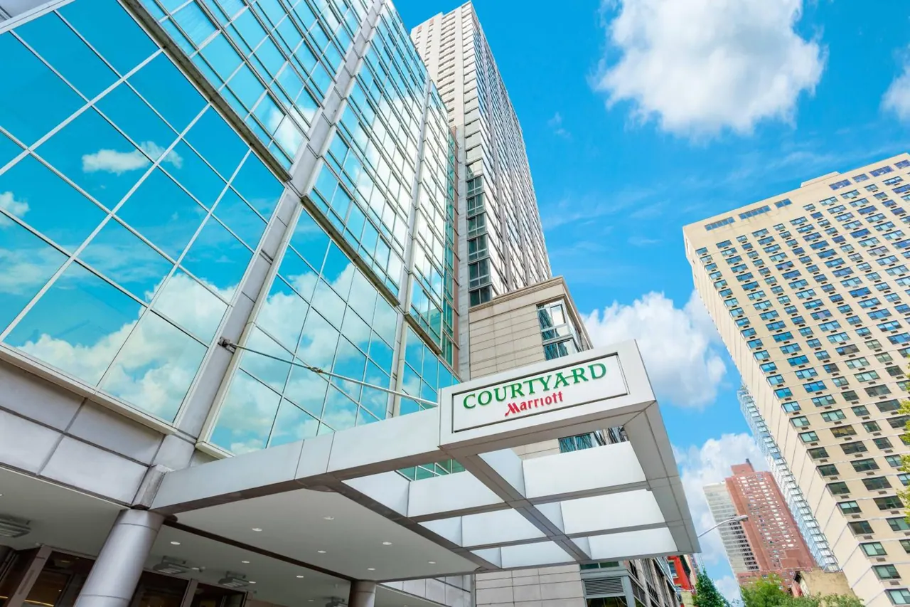 Low-angle view of the Courtyard by Marriott entrance canopy and sign beneath a glass high-rise and neighbouring towers under a blue sky with clouds.