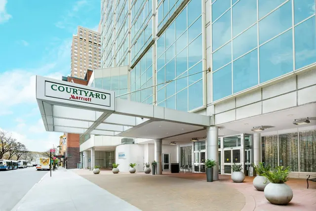The entrance of a Courtyard by Marriott hotel with a glass facade, a canopy sign, and concrete planters under a partly cloudy sky.