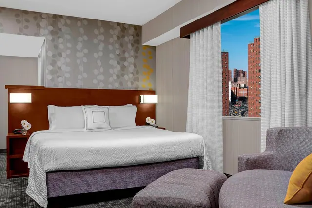 A bedroom with a king bed, an upholstered armchair and ottoman, and a window looking out onto brick apartment buildings and a street.