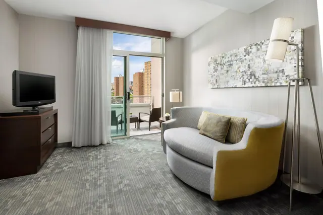A living area with a curved grey and yellow sofa, a flat-screen TV on a wooden dresser, and sliding doors opening onto a balcony with a view of apartment buildings.