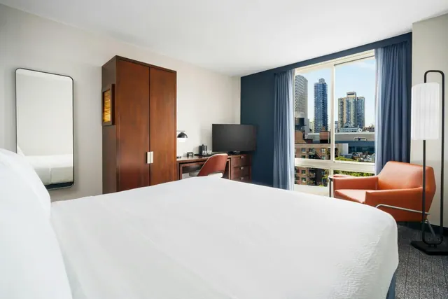 A hotel bedroom with a made-up white king bed, a wooden wardrobe, a desk with a chair and wall-mounted television, an orange armchair, and a large window overlooking high-rise buildings.