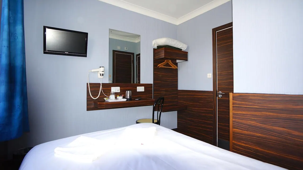 A hotel bedroom with a white bed, wall-mounted television, mirror, wooden desk with a kettle and glasses, a chair, and a wooden cabinet holding folded blankets and hangers.