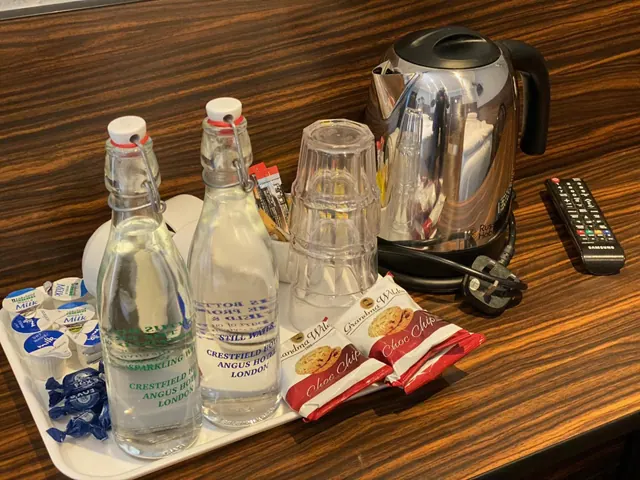 A white tray on a wooden surface holds two glass bottles of water, milk pots, biscuits and a stack of glasses, with a steel kettle and a remote control beside it.