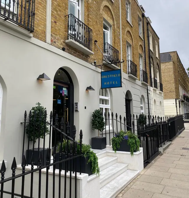 The front of the Crestfield Hotel, showing a white-painted ground floor with a blue hanging sign, black railings and steps, beneath a yellow brick terraced building.