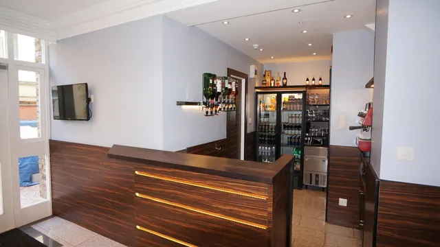 A wood-panelled bar counter with LED strip lighting, with shelves of bottles and a glass-door fridge behind it, a wall-mounted television on the left, and an espresso machine on the right.