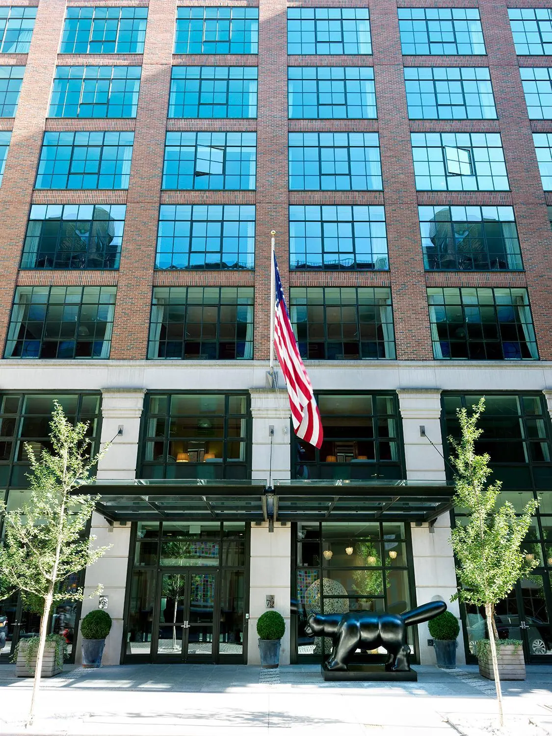 The front of a brick and glass building with a glass entrance canopy, an American flag, a black sculpture and young trees on the pavement.