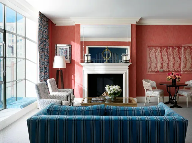 A living room with coral walls, a white fireplace, a blue striped sofa and armchairs, with tall windows showing city buildings outside.