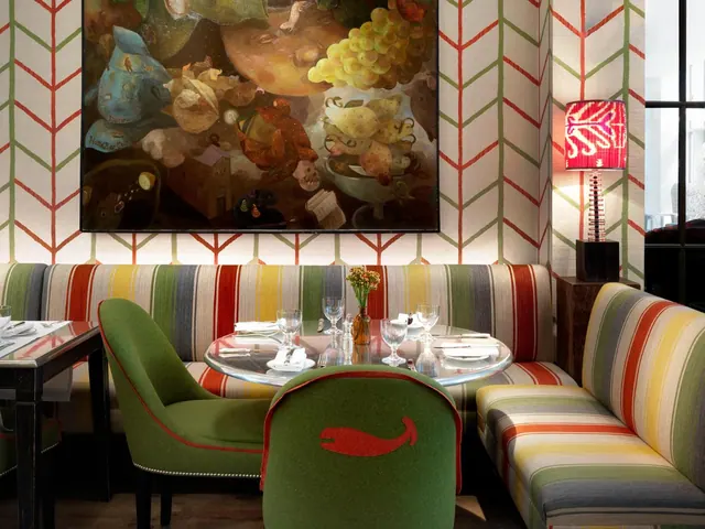 A dining room with a striped banquette, green upholstered chairs, a set round table, a large still life painting and chevron-patterned wallpaper.