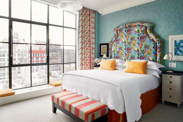 A bedroom with a king bed under a patterned upholstered headboard, a striped bench at the foot, and floor-to-ceiling windows overlooking city buildings.