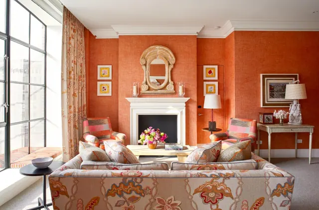A living room with orange walls, a floral-upholstered sofa, two patterned armchairs, a white fireplace and tall windows on the left.