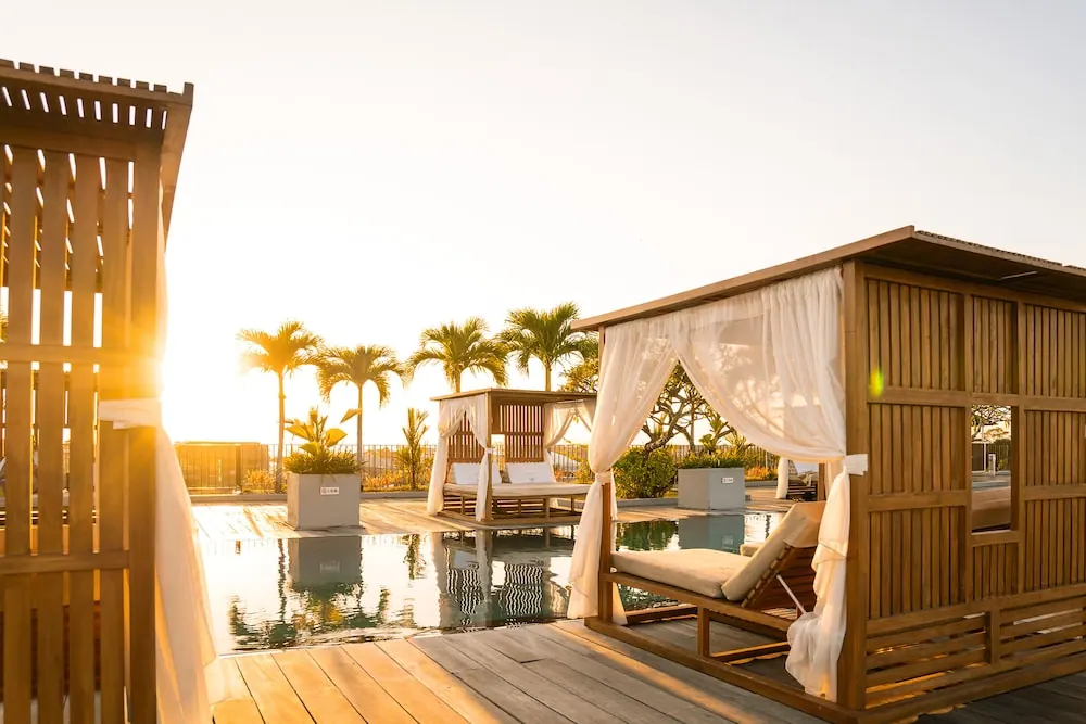 Wooden cabana daybeds with white curtains sit beside a pool at sunset, with palm trees in the background.