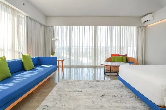 A bright hotel room with a blue sofa, a large bed with white linen, a wicker armchair and a round table, with sheer-curtained floor-to-ceiling windows.