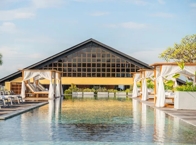 A long pool runs toward a gabled building with a glass front, with white curtained daybeds on timber decks along both sides.