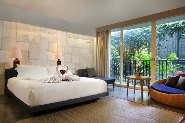 A bedroom with a king bed dressed with a towel swan, a stone accent wall, and floor-to-ceiling glass doors looking onto leafy garden plants.
