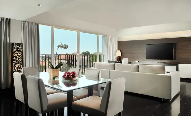 A living and dining area with a glass-topped table, a white sofa, a wall-mounted television and floor-to-ceiling windows opening onto a balcony with rooftops and trees beyond.