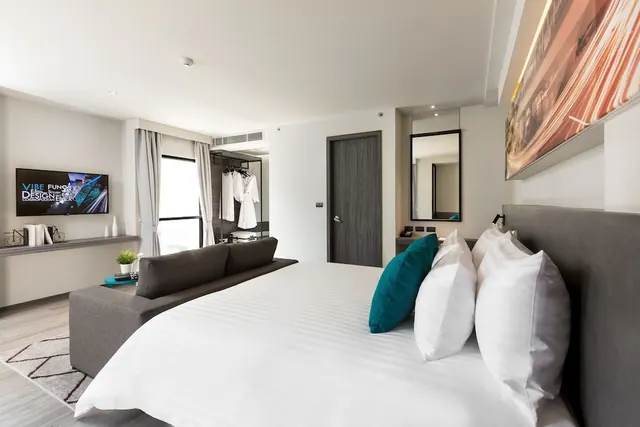 A bedroom with a large white bed, a grey sofa, a wall-mounted television, and an open wardrobe holding two white robes.