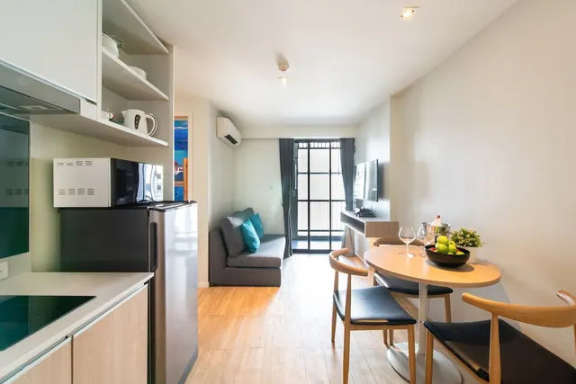 A small apartment-style living area with a kitchenette, grey sofa, wall-mounted television, round dining table with fruit, and a glass door leading to a balcony.