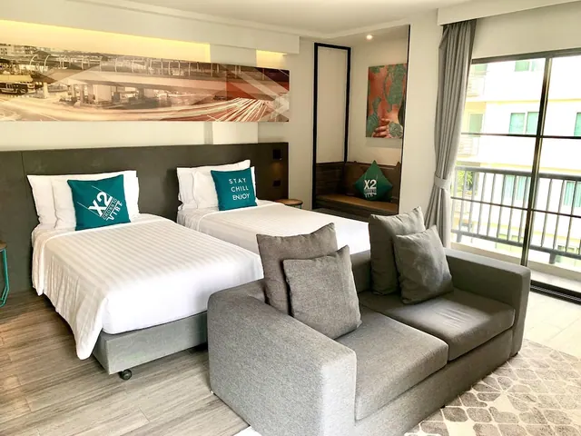 A hotel room with two made-up twin beds with white linen and teal cushions, a grey sofa in front, and a glass door to a balcony looking onto a neighbouring building.
