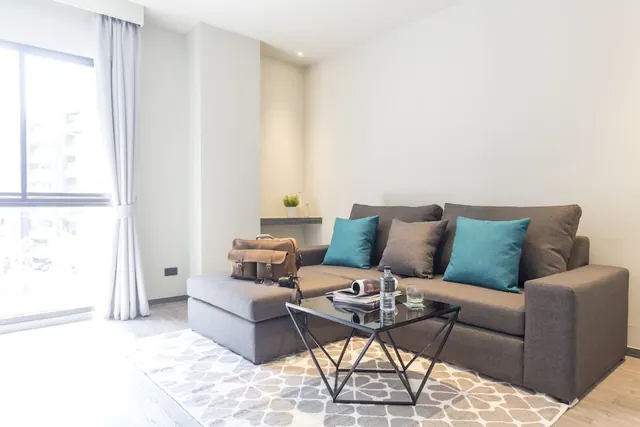 A living area with a grey sofa and teal cushions, a glass coffee table holding a water bottle and magazine, and a window with sheer curtains on the left showing blurred buildings outside.