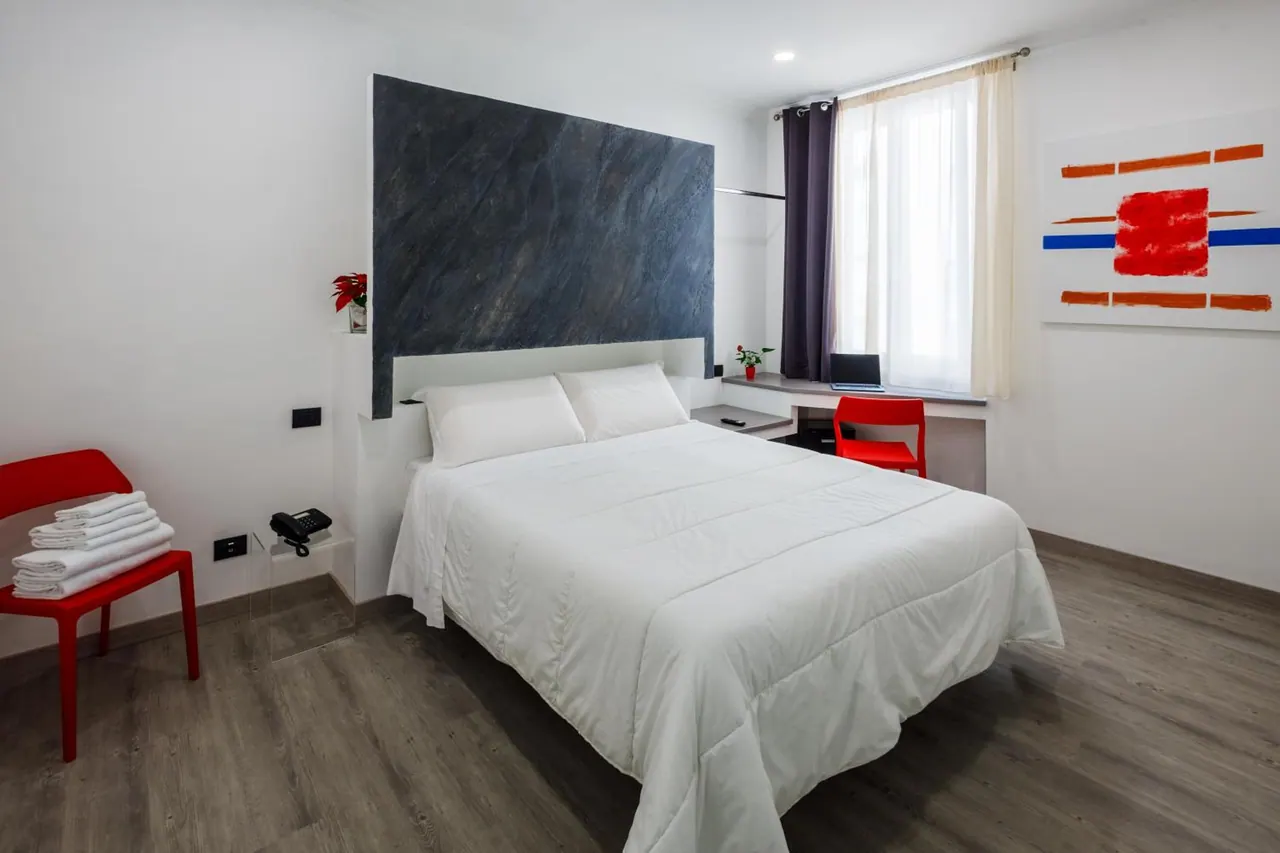 A bedroom with a double bed under a dark slate-look headboard wall, a red chair holding folded towels, a desk by the window and an abstract painting on the wall.