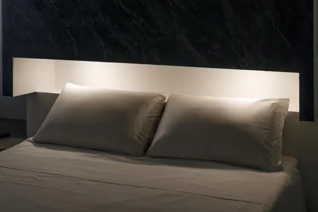 A made double bed with two pillows sits against a backlit recessed headboard below a dark textured wall.