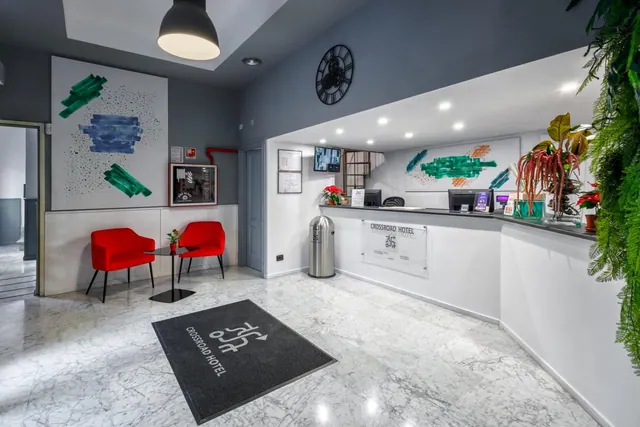 The lobby has a marble floor, a black entrance mat reading Crossroad Hotel, two red armchairs by a small round table, and a reception desk with computer monitors.