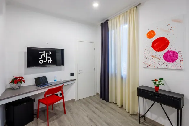 A modern hotel room with a wall-mounted television, a wall desk with a laptop and red chair, a mini fridge, floor-length curtains and an abstract painting.