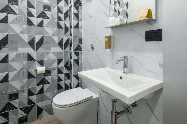 A bathroom with black and white geometric tiles on one wall, a wall-hung basin with a chrome tap, a toilet, and a toilet roll holder.