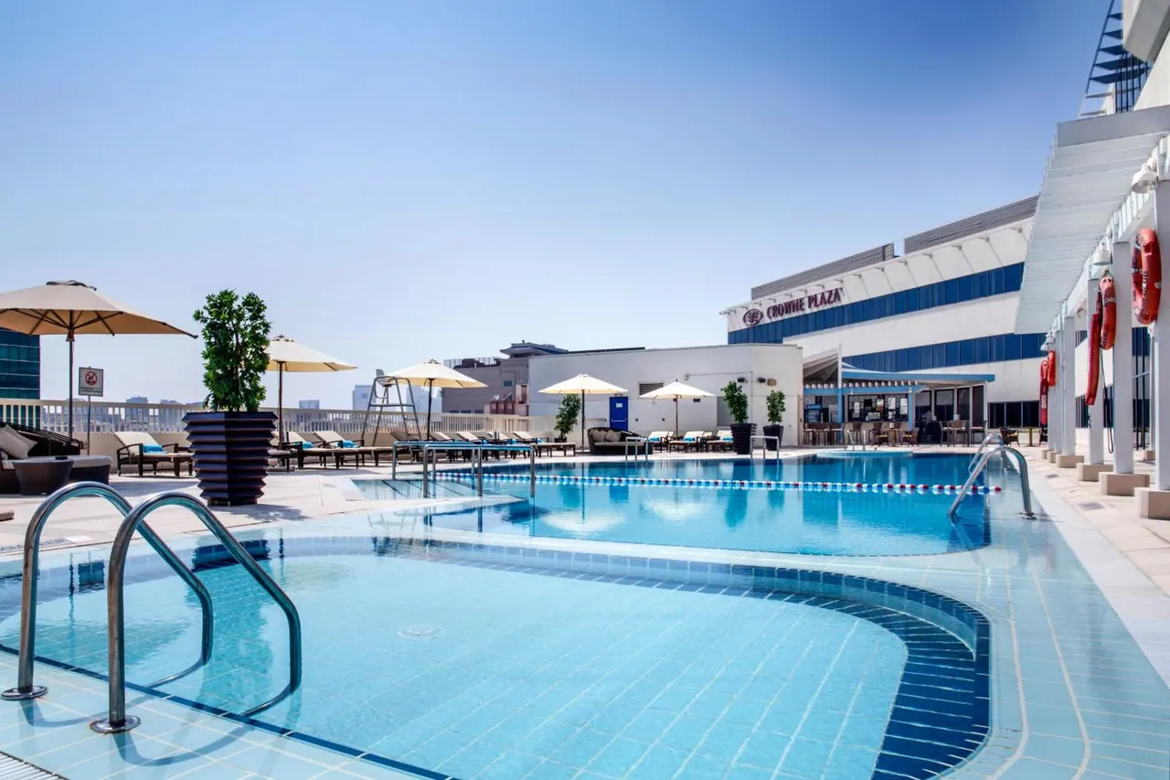 A rooftop swimming pool with lane ropes, sun loungers and parasol umbrellas beside a white building with a Crowne Plaza sign.
