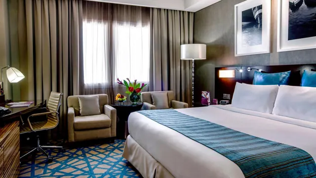 A king bed with blue accent cushions and a teal runner sits beside an armchair seating area and a wooden work desk in a hotel bedroom.