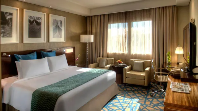 A hotel bedroom with a king bed, two armchairs by the window, a work desk with a lamp, and a wall-mounted television.