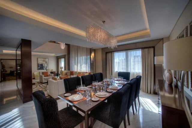 A set dining table with a crystal chandelier overhead, with a sofa lounge area and large windows with sheer curtains visible beyond.