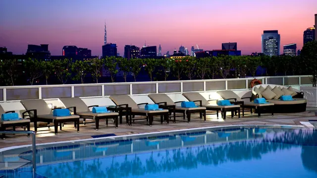 A rooftop pool at dusk with loungers topped with blue towels, and a city skyline with the Burj Khalifa in the background.