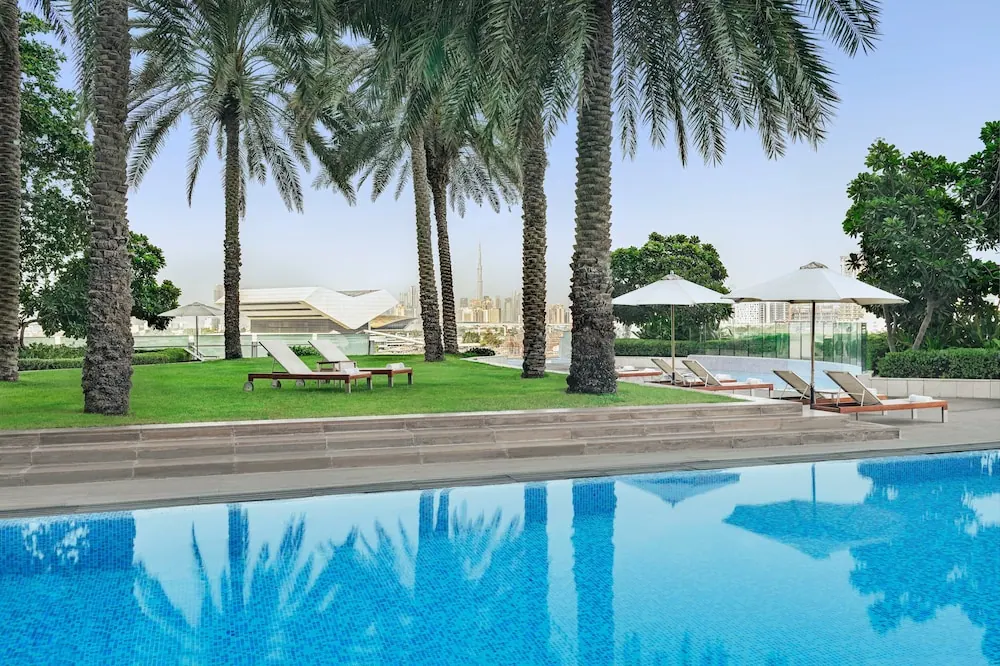 A large swimming pool in the foreground with sun loungers, umbrellas and palm trees, with a city skyline and a tall tower in the distance.