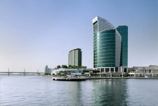 Exterior view of a curved glass hotel tower beside a canal, with a smaller tower and a cable-stayed bridge in the background.