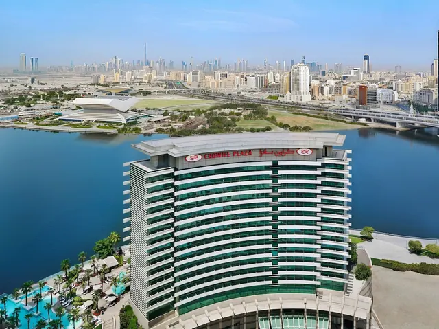 Aerial view of a curved Crowne Plaza hotel tower beside a water canal, with a pool and palm trees at its base and the Dubai skyline in the background.