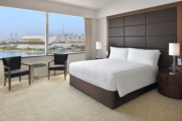 A king bed with white linen faces a wall of windows showing a city skyline, a body of water and a distant tall tower.