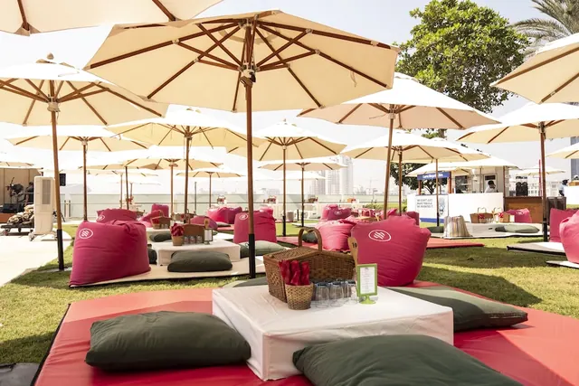 An outdoor lawn seating area with beige parasol umbrellas, pink bean bags, floor cushions on red mats, and a white table holding a picnic basket and glasses.