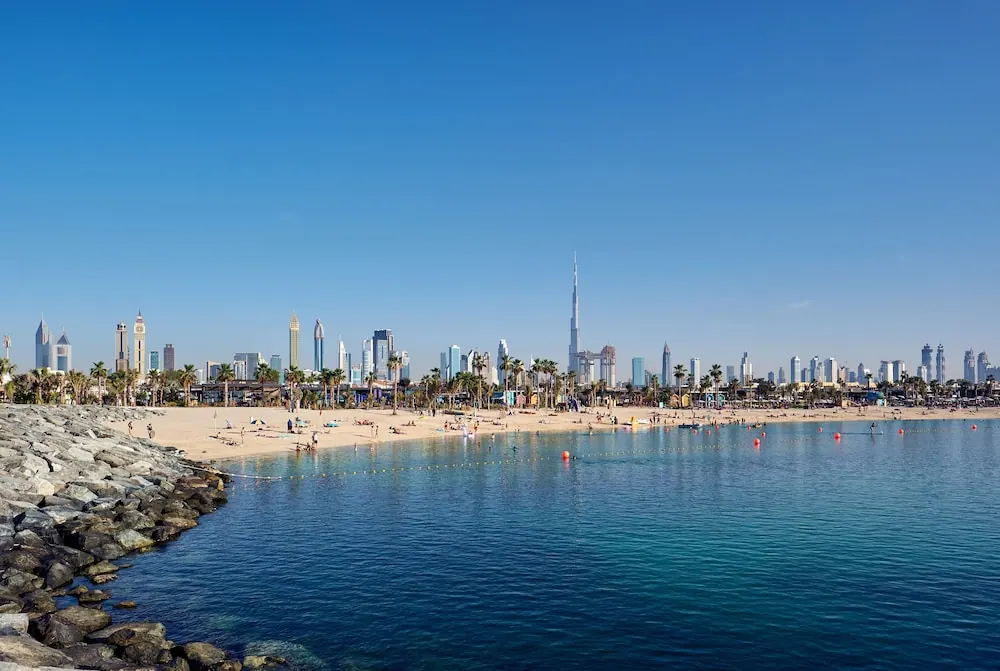A sandy beach with a few people and palm trees runs along calm blue water, with a rocky breakwater on the left and the Dubai skyline, including the Burj Khalifa, in the background.