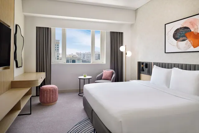 A hotel bedroom with a king bed, a wall-mounted television, a wooden desk, a grey armchair and a window looking out over city buildings.