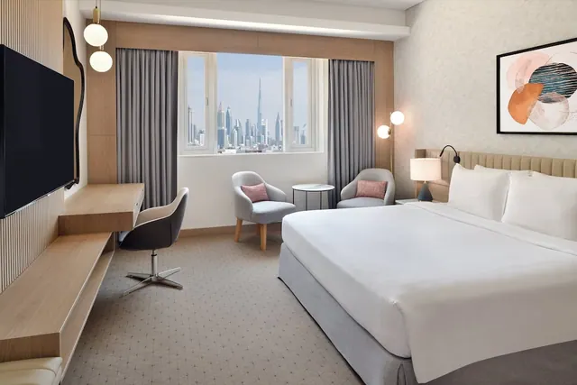 A bedroom with a king bed, two armchairs, a work desk and a wall-mounted television, with a window showing city towers including the Burj Khalifa.