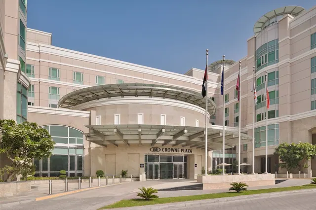 The entrance of a Crowne Plaza hotel with a curved glass canopy, several flags on poles, and planters under a clear blue sky.