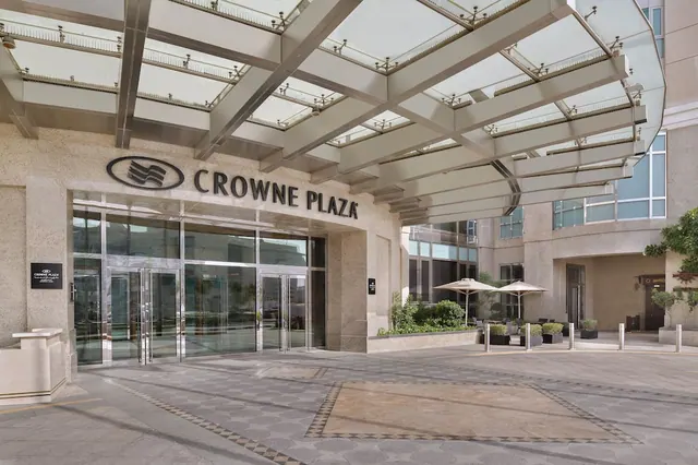 Hotel entrance facade with the Crowne Plaza sign, a glass canopy overhead and a patterned stone forecourt, with no people visible.