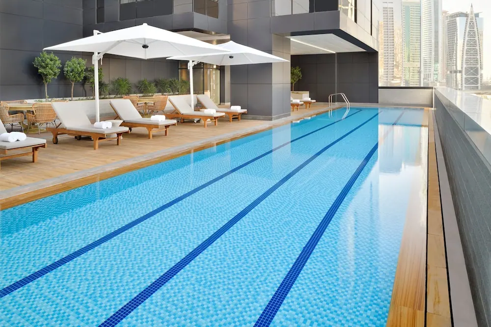 An outdoor lap pool with blue tiled lane lines is flanked by white sun loungers, white umbrellas and a wooden deck, with a city skyline of tall towers visible beyond the railing.