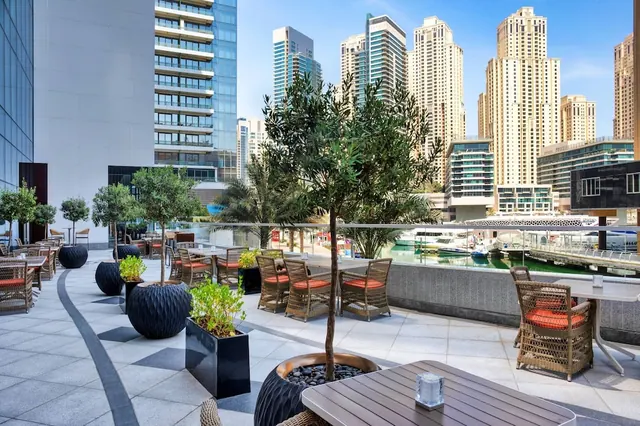 An outdoor terrace with olive trees in planters, wicker dining chairs with red cushions, and high-rise towers and a marina with yachts behind it.