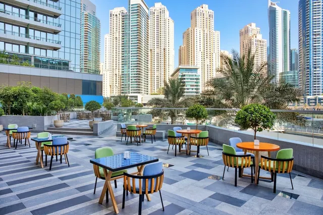 An outdoor terrace with green and blue chairs and round tables overlooks a row of high-rise towers and palm trees under a clear blue sky.
