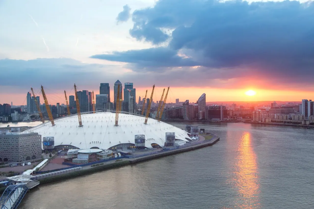 Elevated view of The O2 dome beside the River Thames, with Canary Wharf towers and the city skyline behind it at sunset.